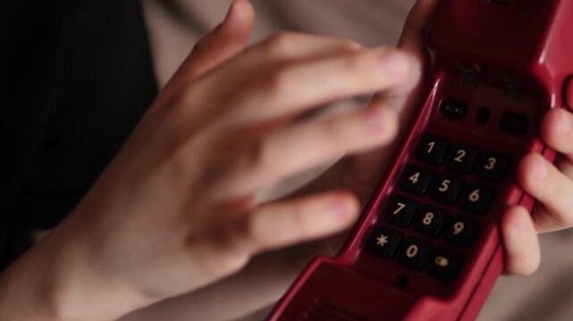 A close-up of a child's hands pressing the buttons on the keypad of a red retro telephone. The boy dials a number on the vintage receiver, getting ready to make a phone call.