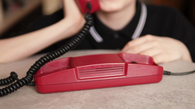 A close-up of a child's hand picking up the receiver of a stationary red retro telephone. The boy lifts the vintage device handset from the table to start a conversation.