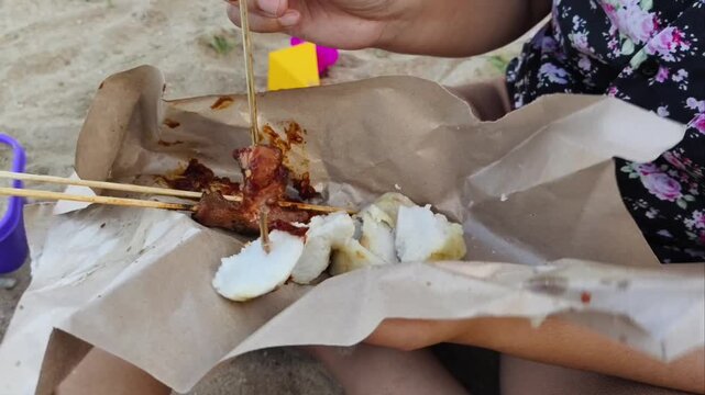 Eating traditional Indonesian chicken satay with rice cakes on a sandy beach.