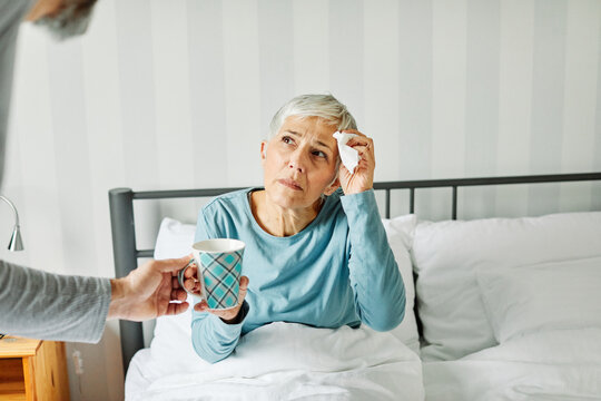 Portrait of a sick senior elderly woman sneezing and using tisue in bed in the morning, a mature woman at home, cold or flu illness and pain, senior couple, husband offering hot tea to his wife, love 