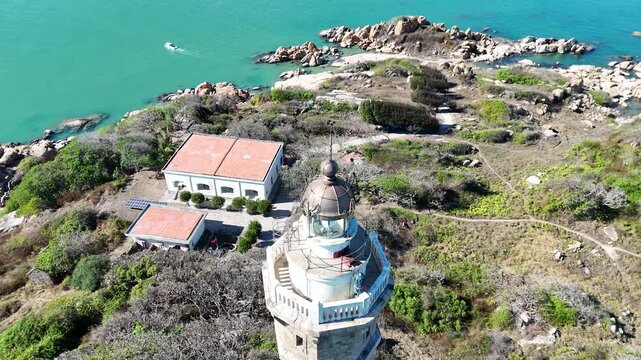 Drone orbit around the top of Ke Ga Lighthouse revealing the historic beacon, rocky island and turquoise waters off the coast of Binh Thuan, Vietnam.