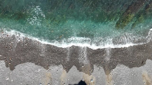 Top down aerial view with movement to the right of turquoise sea waves crashing on a pebble beach coastline