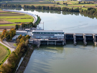 Aerial view captures hydroelectric power plant by a river surrounded by fields and trees. Views show roads and agricultural land nearby. © 24K-Production