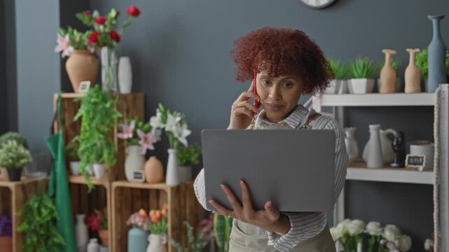 Woman florist holding laptop with hand while talking on smartphone inside building, apron visible and flowers around; small business focused.