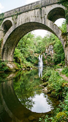 Fototapeta premium Bajo los arcos de piedra de un puente antiguo, reflejos en el agua tranquila con vegetación verde y un pequeño salto de agua al fondo.