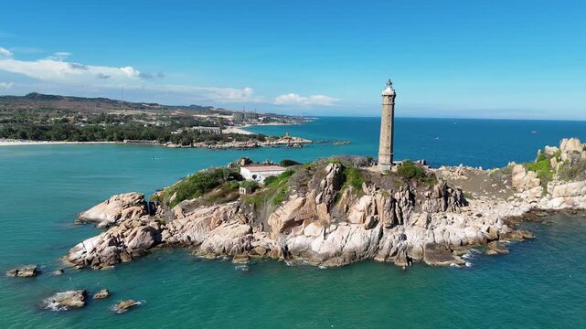 Cinematic aerial flyby of Ke Ga Lighthouse rising above granite rocks and crystal clear coastal waters in Binh Thuan Province, Vietnam.