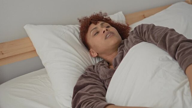 Woman covering ears for earache while clutching white duvet on wooden bedframe in building; pain exhaustion.