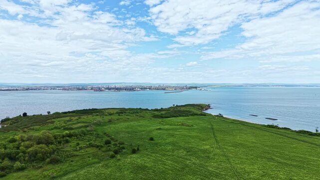 A view shows a green land patch next to a large body of water with a city in the distance under a partly cloudy sky. The scene captures nature and urban life together.