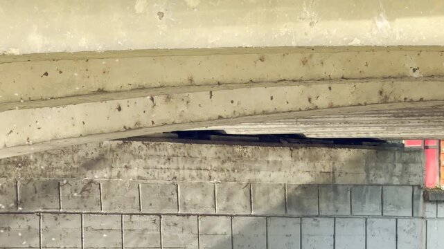 Weathered concrete bridge beams span securely over a structured block retaining wall. The gritty texture and stark shadows evoke a raw, industrial mood of urban endurance.