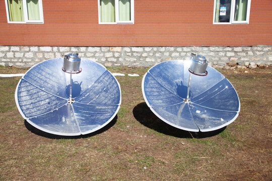 Solar cookers positioned in a grassy area outside a stone house in the Himalayan region of Nepal, showcasing modern sustainable technology against a backdrop of mountains and clear blue sky