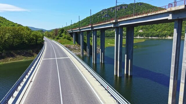 A road runs parallel to a river with a bridge crossing over it. The landscape features green hills and clear blue skies with no clouds. It is daytime and sunny.