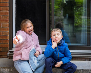 Cute smiling little blond girl and boy fooling around, sticking their tongue out and doing a thumb up while sitting outdoors, Montreal, Quebec, Canada © Anne Richard