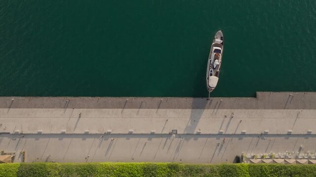 Aerial view of the Island of Ortigia waterfront featuring a docked boat, people walking on the promenade, and a green hedge by the sea in Syracuse, Sicily, Italy.