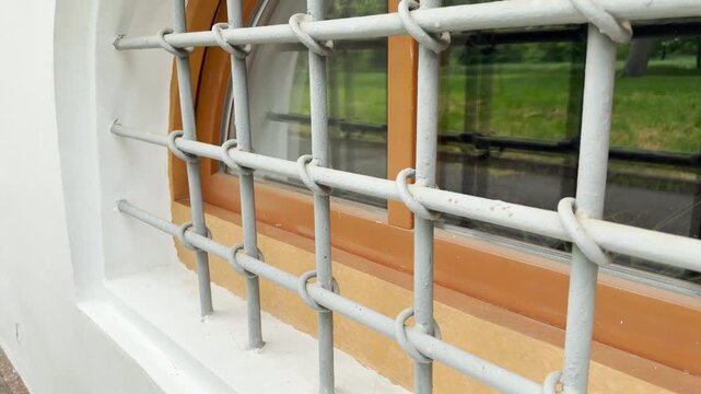 A sturdy metal security grille sits over a wooden-framed glass window on a clean white wall. The structured bars offer a strong feeling of safety, protection, and quiet stability.