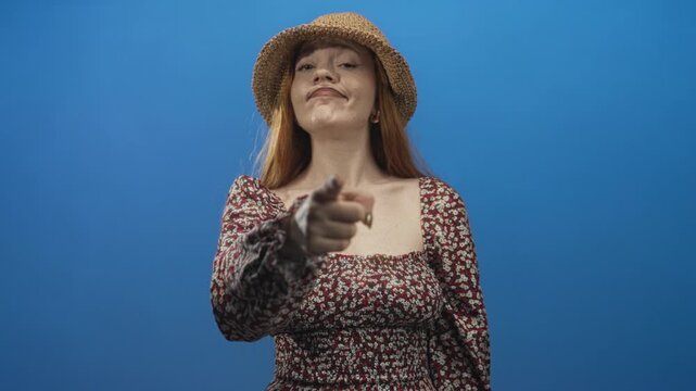 Woman points finger at camera in studio wearing straw hat and floral dress, smirking toward lens; playful defiance.