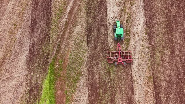 A tractor is in a large field working to plow the ground. The tractor pulls a plow as it moves along the soil. Green areas show growth while other parts are bare. The sky is clear and bright.