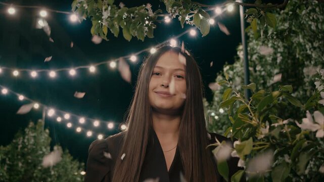 Woman stands in park under festoon lights and falling petals, looking directly into camera with gentle smile. Blooming trees and dark background create dreamy evening portrait.