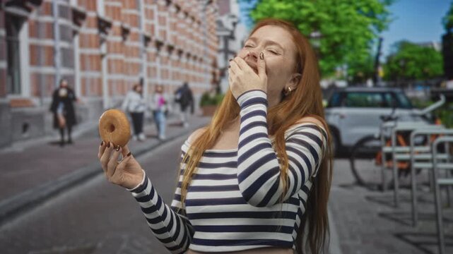 Redhead woman holding donut with hand to mouth laughing on urban street, striped top visible; carefree joy.