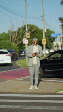 Static shot of mature Brazilian executive in gray plaid suit checking smartphone at urban crosswalk with moving traffic