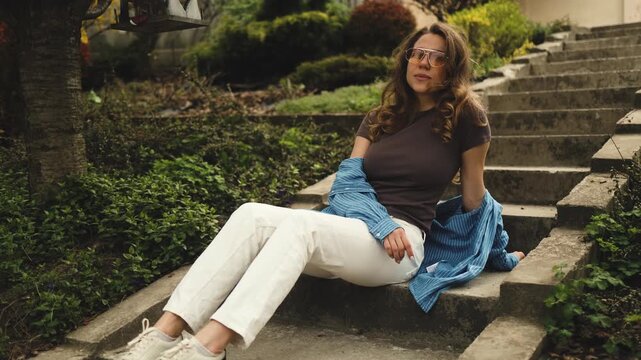 A stylish young woman sits casually on outdoor stone steps in a green garden or backyard. Female portrait, fashion model look, urban landscape, relaxed lifestyle, summer or spring street style