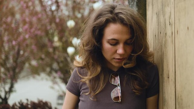 A portrait of a thoughtful young woman leaning against a wooden fence with her eyes cast down. Pensive look, melancholy mood, female reflection, casual lifestyle, blooming spring garden background