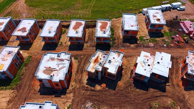 High angle view of unfinished brick houses on construction site. Aerial view of rows of residential houses under construction with concrete roofs and dirt pathways.