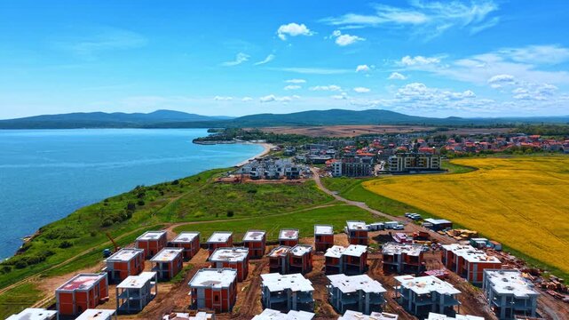 Scenic aerial panorama of seaside cottage development under construction. Aerial view of a new residential resort village being built by the blue sea and yellow flower field.