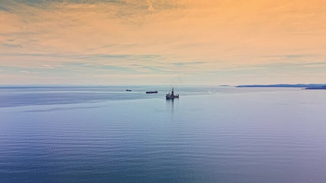 An oil tanker travels through the open sea under a clear sky. In the background, there is another vessel and a drilling rig. The scene shows typical maritime activity.