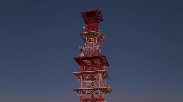 Tall telecommunications tower with red and white metal structure featuring multiple satellite dishes and antennas against deep blue twilight sky. Red warning lights blink on the lattice framework.