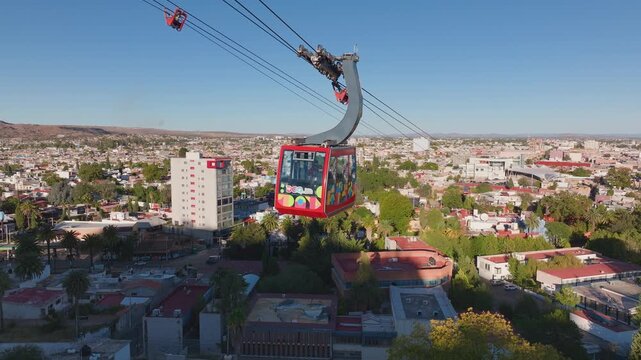Aerial view of vibrant red cable car gondola with colorful graphics gliding over urban landscape. Palm trees and residential buildings spread across hillside terrain under clear blue sky.