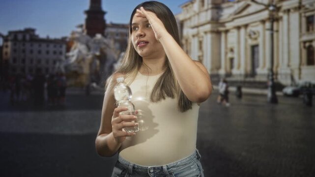 Young woman holds water bottle and shades eyes with hand while standing in a sunlit building square near ornate fountain and crowd; relief.