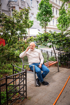 Smiling mature man sitting alone on city park bench with smartphone and backpack