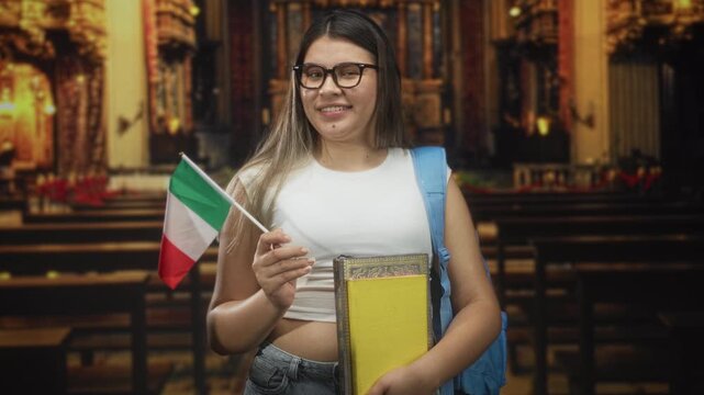 Young woman holding italian flag and stack of books, smiling with backpack visible inside ornate building; study abroad pride.