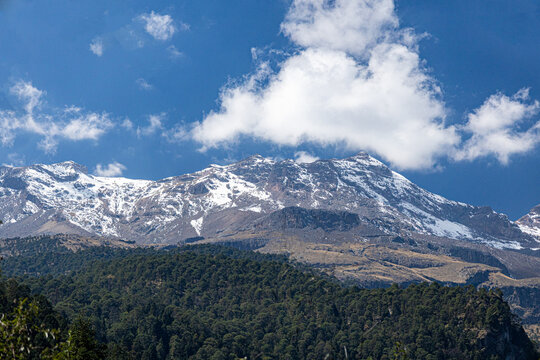 Volc&aacute;n Iztacc&iacute;huatl nevado sobre un denso bosque de pinos bajo un cielo azul intenso con nubes blancas, M&eacute;xico