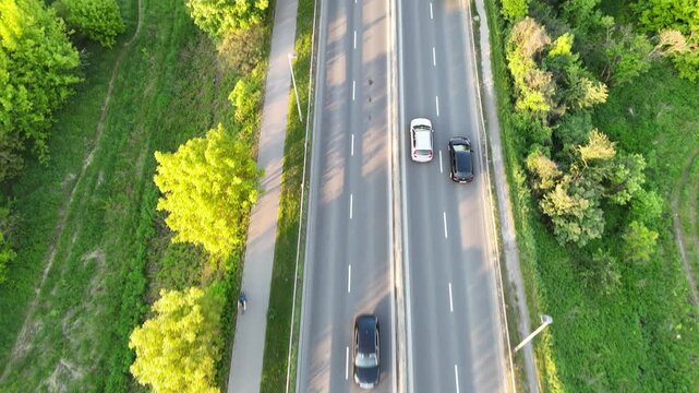 Aerial view of vehicles traveling along a tree-lined road, showcasing the continuous flow of traffic amidst lush greenery and urban elements in a suburban setting. Cars riding at rural route