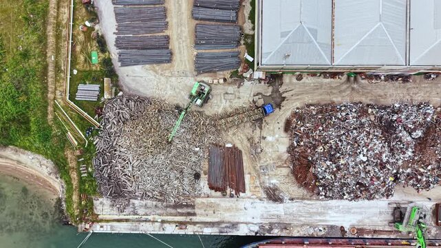 Workers use equipment to move and sort materials at a construction site next to water in a busy industrial zone during the day. The area shows piles of wood and metal scraps.