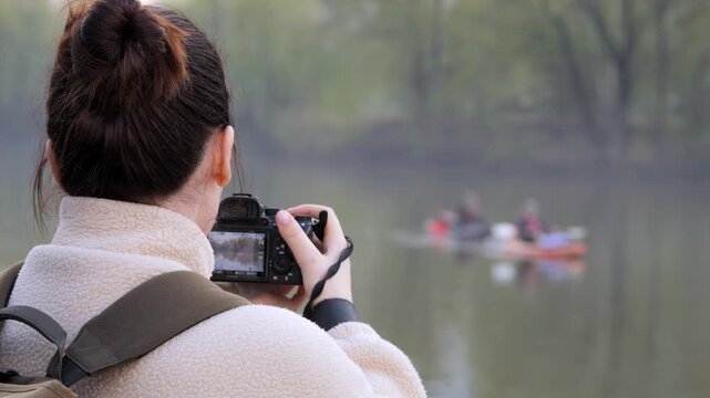 Woman holds camera, focuses on people in canoe on calm lake. Trees line shore, fog softens background. She wears white coat, backpack. Scene peaceful, natural light. No animals or plants named