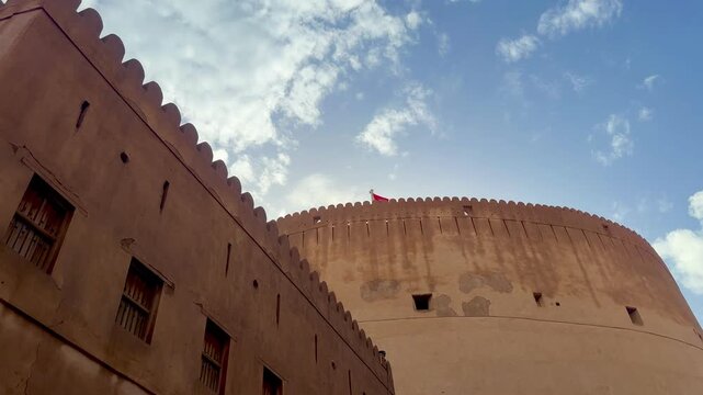 Historic Nizwa Fort after rain under clear blue sky with Omani flag and mosque minaret in background. Nizwa, Oman - May 14 2026