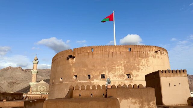 Historic Nizwa Fort after rain under clear blue sky with Omani flag and mosque minaret in background. Nizwa, Oman - May 14 2026