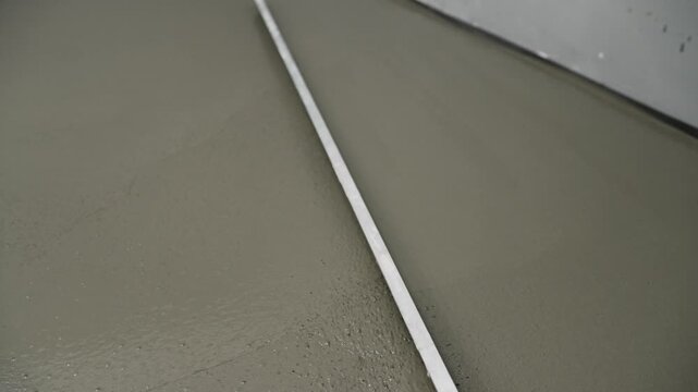 Close up of worker using long trowel to spread and level wet self leveling screed on concrete floor. Process of creating a smooth surface for new flooring installation