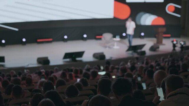 Audience watching keynote presentation at busy tech conference, speaker on stage, large screen visuals, attendees using phones and laptops, dim auditorium lighting.