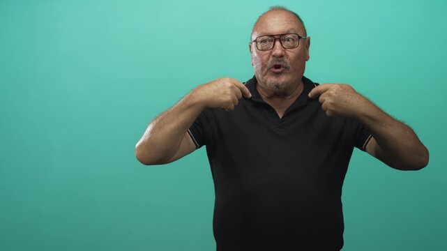Man pointing fingers to chest wearing glasses and black polo against teal backdrop in studio; surprise confidence.
