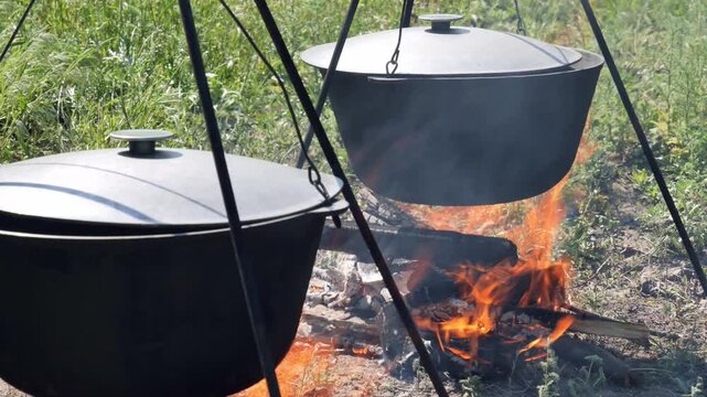 Close-up shot of traditional cast-iron cauldrons (kazan) boiling over a bright campfire wood fire. Thick smoke and dancing flames under the pots