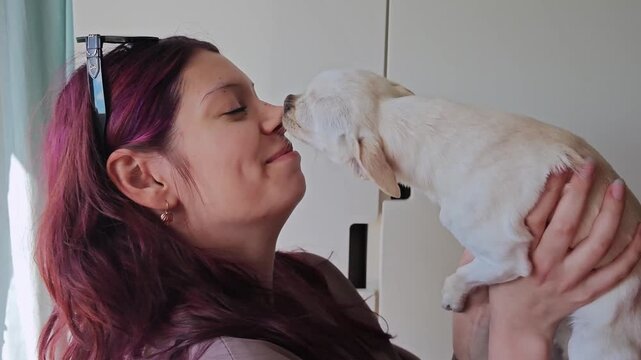 Woman with purple hair kisses small white dog in a bright indoor setting, showcasing affection and connection between pet and owner in a cozy environment
