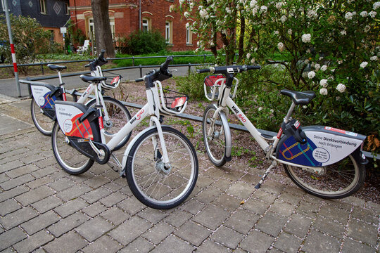 VAG Rad rental bicycles parked in a row on a paved sidewalk next to green flowering bushes.