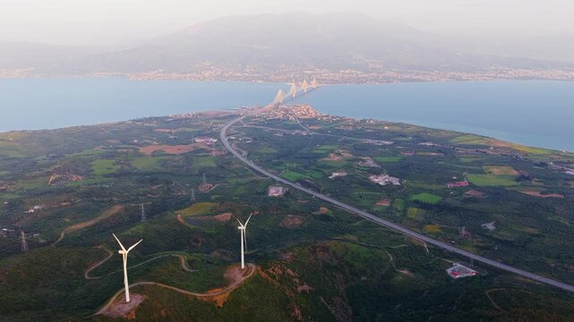Stunning aerial panorama of wind turbines on a green coastal hill, with the magnificent rio-antirrio bridge spanning the gulf of corinth in the background under a soft, hazy sky