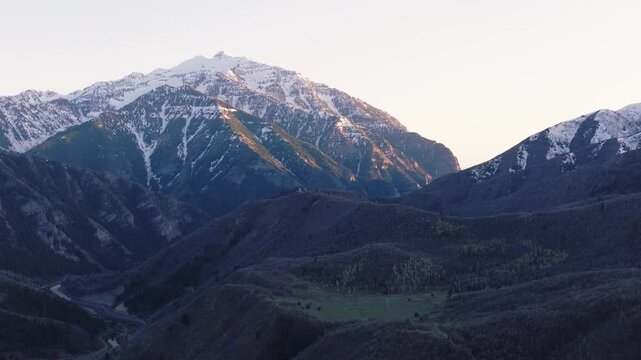 Aerial view of snow-capped mountain peaks in Provo Canyon with golden hour light hitting the rugged slopes and dark forested valleys in Orem, Utah, United States.