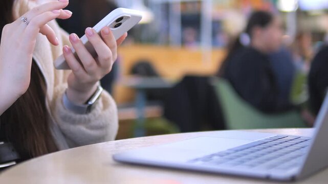 Close-up of a person using a white smartphone at a table. Laptop sits open beside them. Background shows blurred people in a casual indoor space. Hands move across the phone screen with focus