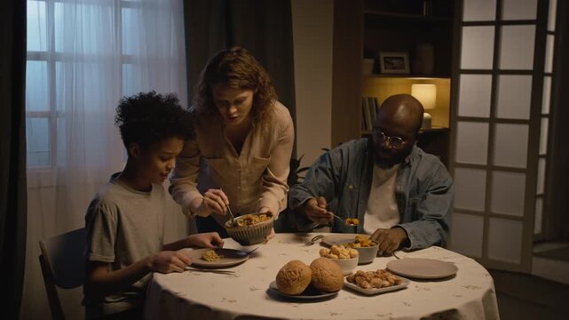 Cozy lifestyle scene of multiracial family enjoying dinner together at home with caring mother serving food to son and husband, copy space