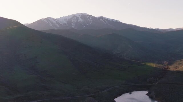 Aerial view of Provo Canyon featuring snow-capped mountain peaks, lush green hills, and a calm lake during the soft light of dawn in Orem, Utah, United States.